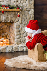 A small child in a red Santa Claus costume, plays in his residence, licks a New Year's ball like candy. Festive postcard. The blond boy laughs. Christmas tree with boxes by the festive stone fireplace