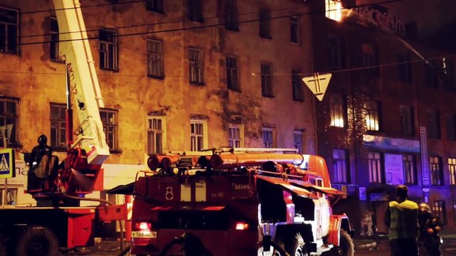 Firefighters with fire hose and fire engines or trucks are fighting fire in residential building at night. Huge smoke, hot flame in burning house
