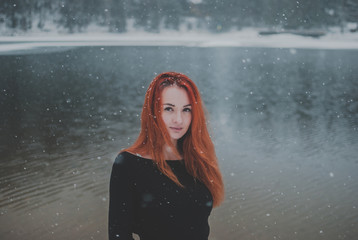 Joyful handsome black-eyed redhead girl looking at the camera. Splendid shady black mountain lake reflecting trees of forest behind her back.