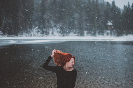 Handsome redhead girl looking in the distance and playing with her gair. Snowflakes falling on her head. Splendid shady black mountain lake.