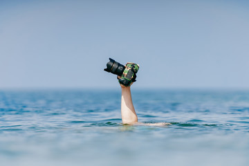 Unrecognizable male hand holding expensive professional photocamera in waterproof military case above water in ocean.  Odd photographer working in extreme dangerous conditions. Safety and protection.