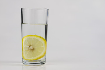 A glass of cold water with lemon on a white kitchen table. A refreshing drink in warm weather.