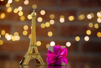 Fototapeta na wymiar handmade gift box with purple bow and Eiffel tower golden souvenir on wooden table with fairy lights on bokeh background