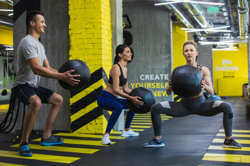 Energetic man with girlfriend are doing squats with heavy medical ball. Fit female coach is showing exercises to them while using same equipment. They are straining wide plie sit-ups for leg and