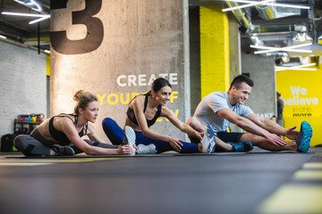 Positive two women and man are doing flexibility exercises together in fitness center. They are sitting on one hip and straightening other leg while bending body to it. Class exercising simultaneously