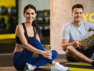 Focus on smiling lady sitting on floor during training in sport studio. Positive man is having good time while being near her and holding outfit. They are having workout indoors with foam rollers