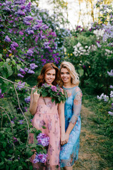 Romantic mood. Twin sisters tender relationship. beautiful twins young models in colorful summer dresses posing for camera in blooming lilac garden. Cheerful cute stylish girls in magic park outdoor.