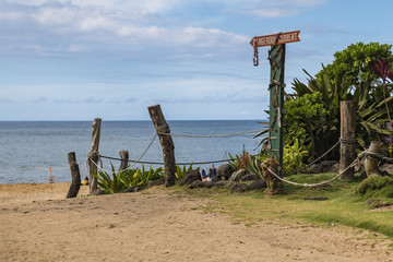 The entrance to Waimea Beach in the north shore of Oahu, Hawaii.  There is a "Dangerous current" sign full of leis.