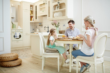 Positive beautiful young family in casual clothing sitting at dining table and chatting while having breakfast in morning