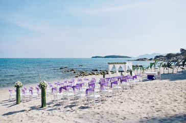 Fototapeta na wymiar Beach wedding venue settings on the white sand with white chiavari chairs decoration with purple organza sash, minimal flower decoration at seaside with panoramic blue sea on sunny day before sunset