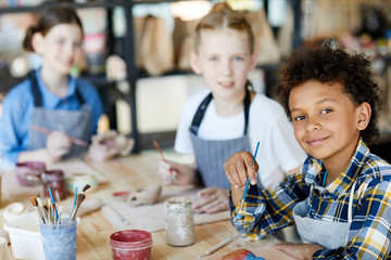 Happy mixed-race boy with paintbrush looking at you at lesson of art and craft