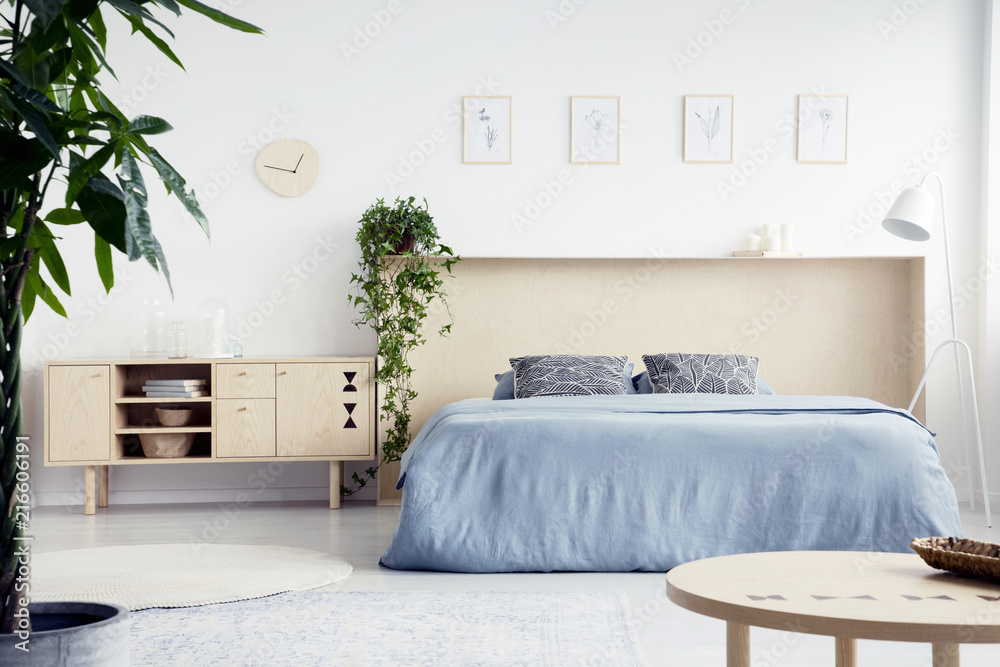 Wall mural Real photo of white bedroom interior with double bed with blue sheets and box bedhead, fresh plants and cupboard with books and glass vases