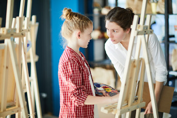 Little girl with color palette listening to her teacher while both standing by easel at lesson of painting