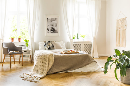 Breakfast tray on a comfy bed with cozy beige sheets and blanket in a stylish, white bedroom interior with a modern gray armchair