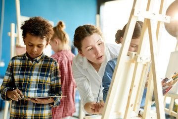 Young teacher standing behind one of pupils and consulting her about painting technique