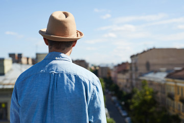 Rear view of young man in hat and casual shirt contemplating cityscape while standing on roof and thinking about life