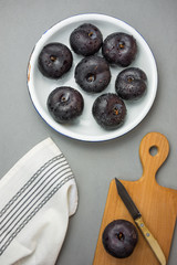 Dark red big plums on white vintage enamel plate. Wooden cutting board knife white cotton towel on gray stone background. Minimalist style. Autumn fall harvest. Top view