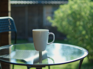 Cup of coffee on glass table with backdrop of sun light, grenn leafs, trees and house. Summer morning, selective focus.