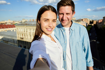 Selfie of positive beautiful young couple in casual shirts smiling at camera while standing on roof in city, tourists concept