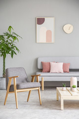 Grey wooden armchair next to table in living room interior with poster above couch. Real photo