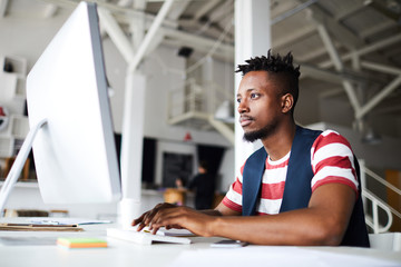 Fototapeta premium Young serious businessman concentrating on network while sitting by desk in front of computer monitor