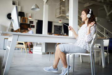 Young relaxed businesswoman in casualwear listening to music in headphones while sitting by desk in office