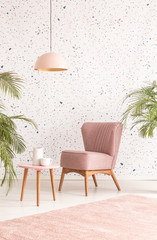 Pink lamp above wooden table and armchair in pastel living room interior with plants. Real photo
