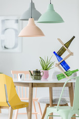 Three pastel lamps, dining table with a wine holder, bottles, plant and pot, and yellow chair in a...