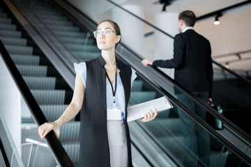 Young female delegate with document moving downwards on escalator after business conference or...