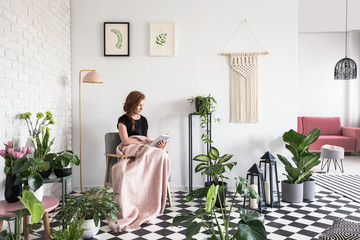 Young girl reading a book, covered with blanket in a living room interior with plants and checkered...