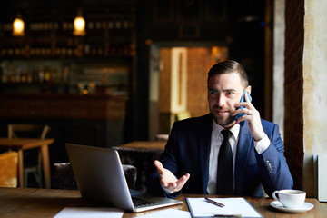 Content confident handsome businessman in formal jacket sitting at restaurant table and solving work issues while talking on mobile phone and gesturing