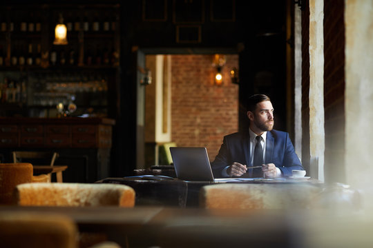 Serious contemplative young businessman in expensive suit sitting at table with laptop and twisting pen while thinking of project and looking out window in loft restaurant.