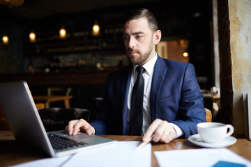 Busy confident handsome male business executive checking online document while using laptop and sitting at table in modern cafe