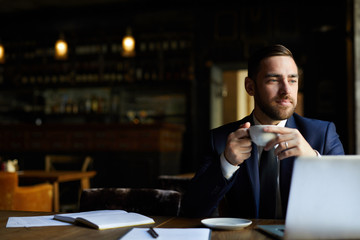 Content dreamy handsome businessman with beard holding coffee cup and looking away while enjoying coffee break in restaurant