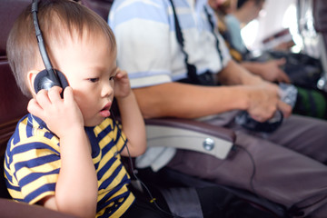 Cute Asian 30 months / 2 years old toddler boy child wearing headphones listening to in flight entertainment on board while traveling in airplane, Flight entertainment for kid, little traveler concept