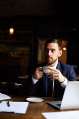Pensive frowning handsome businessman with beard sitting at table with laptop and drinking hot coffee while waiting for service staff in restaurant