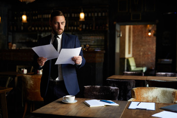 Serious concentrated handsome young restaurant owner in formal suit examining financial papers and...