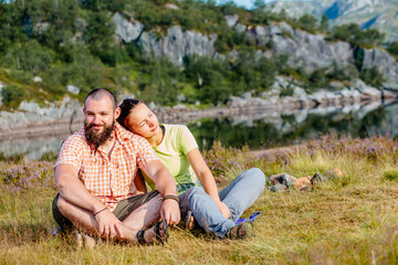 Young couple on nature sitting on the grass with beautiful flowers and lake on background. Travel, vacation, holidays and adventure concept.