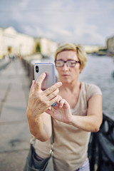 Aged lady in glasses leaning on fence near river and posing for selfie on street of beautiful city