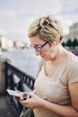 Senior lady in glasses standing on city street near river and using modern smartphone