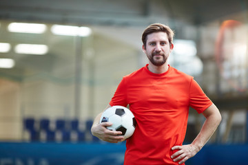 Young football player in red uniform holding soccer ball with the other hand on his waist
