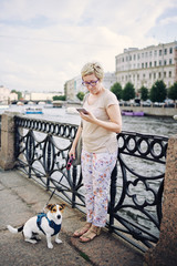Senior female in casual outfit holding adorable dog on leash and looking at smartphone while leaning on fence near river on city street