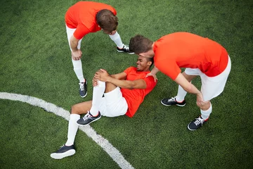 Foto op Plexiglas Two young football players in uniform leaning over their mate lying on field with hurt knee © pressmaster