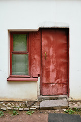 White plaster wall of old house with small red window and crumbling door