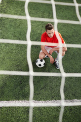 Young football player with ball squatting on green field in front of gates with net