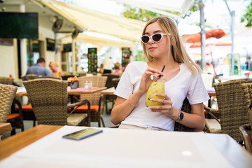 Young beautiful woman in round sunglasses with cocktail at the terrace of cafe having fun