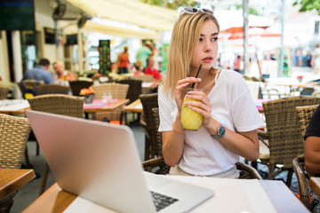 Beautiful smiling young woman sitting outdoors and chatting, drinking cocktail using laptop a in cafe.