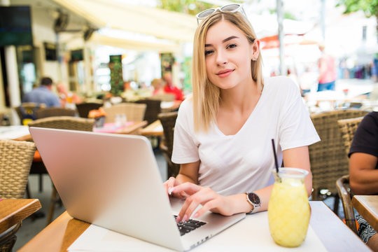 Young woman sits in front of opened laptop computer, drinks fresh summer cocktail, enjoys online communication and free internet