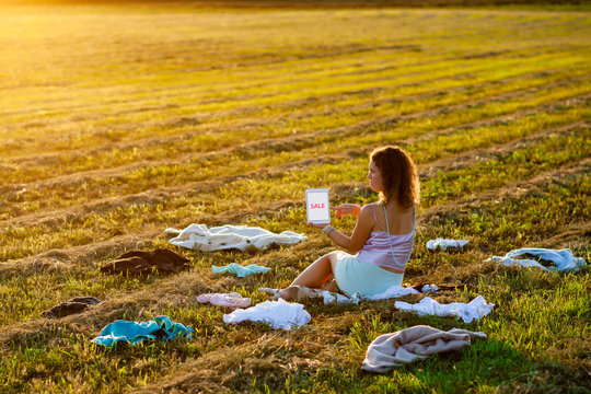 Dressy woman sitting in the middle of the field between scattered cloth and holding a tablet with SALE title during sunset. Fast fashion over-production and pollution concept.
