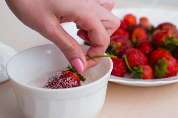 Woman dips the strawberries in the sugar. Close - up woman's hand next to a plate of strawberries.